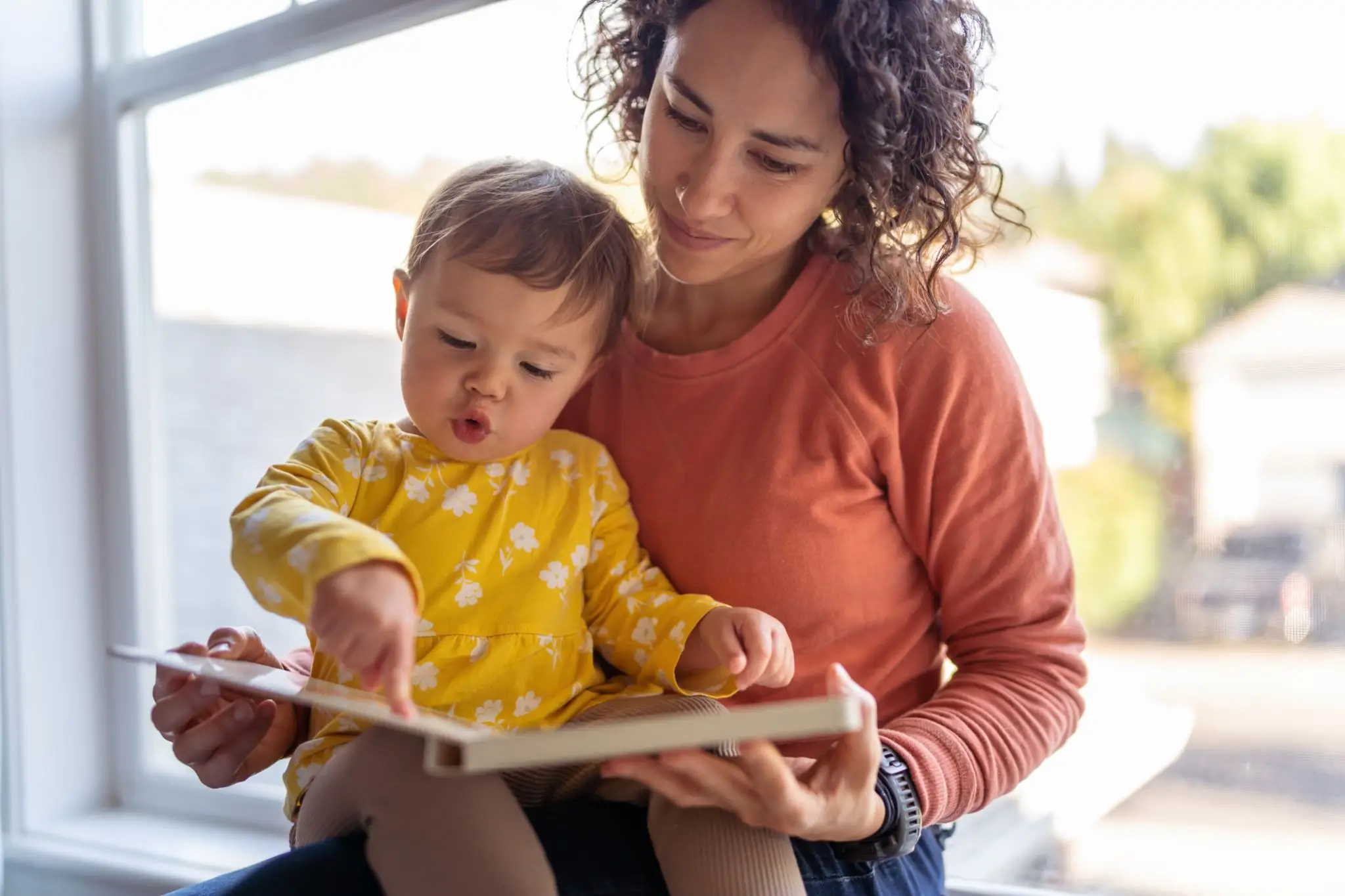 Adult reading book with toddler by window